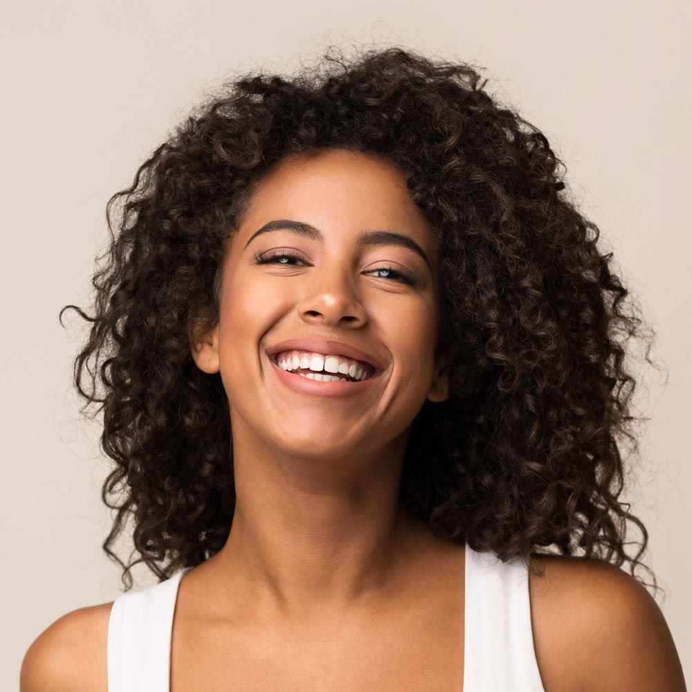 Smiling person with curly hair in a white tank top against a beige background