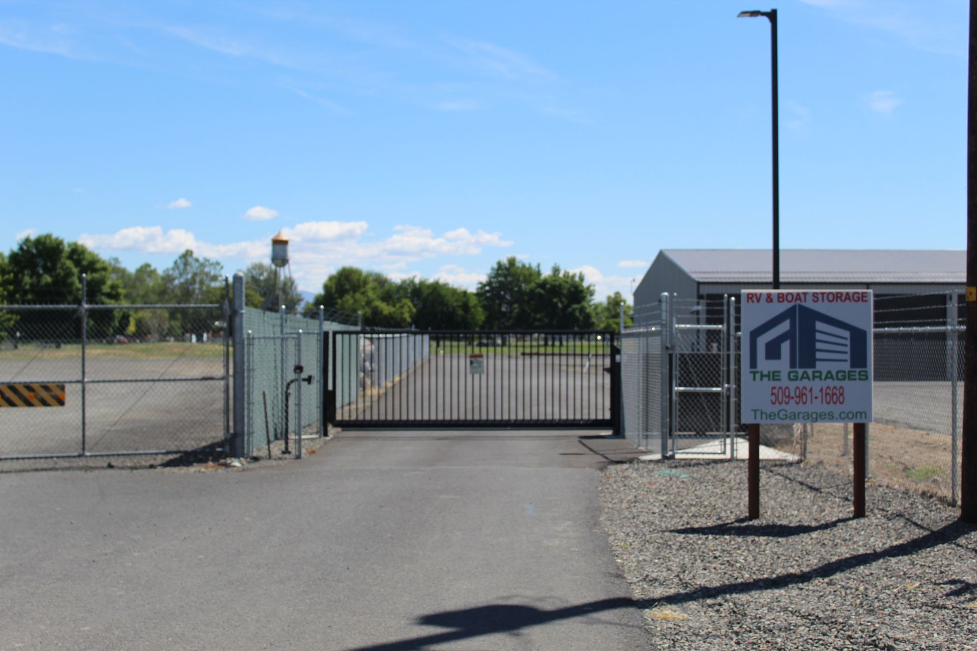 Gated entrance to a gravel lot with a sign reading “The Grand,” under a clear blue sky.