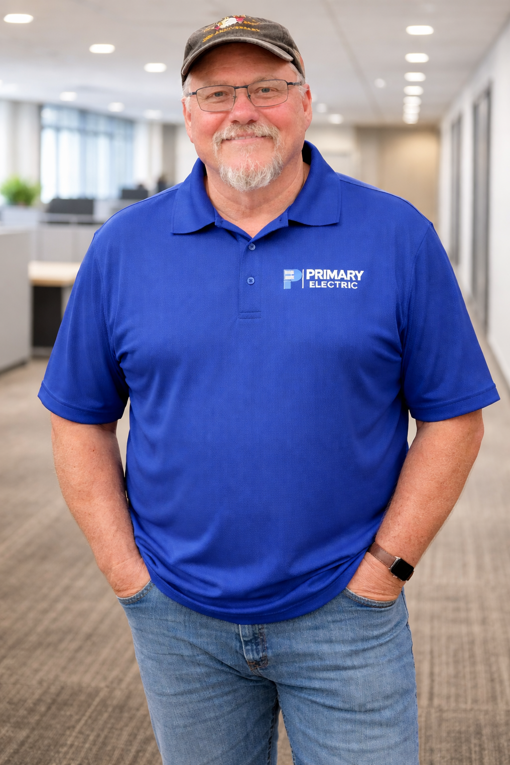 Man in a blue polo and jeans stands smiling in a bright office hallway, wearing a cap and smartwatch.