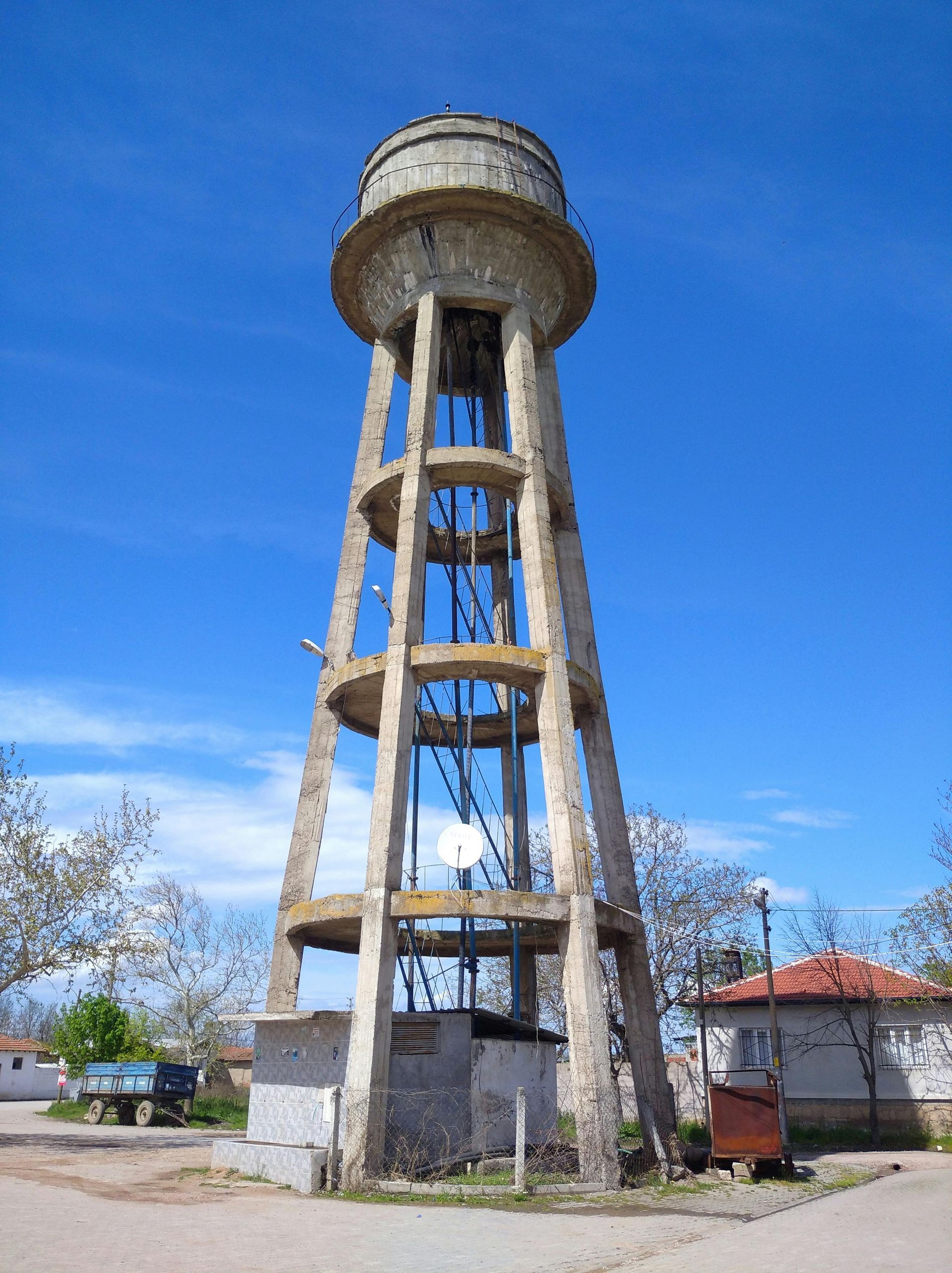 Tall white water tower with blue supports against a clear blue sky in a small park-like area
