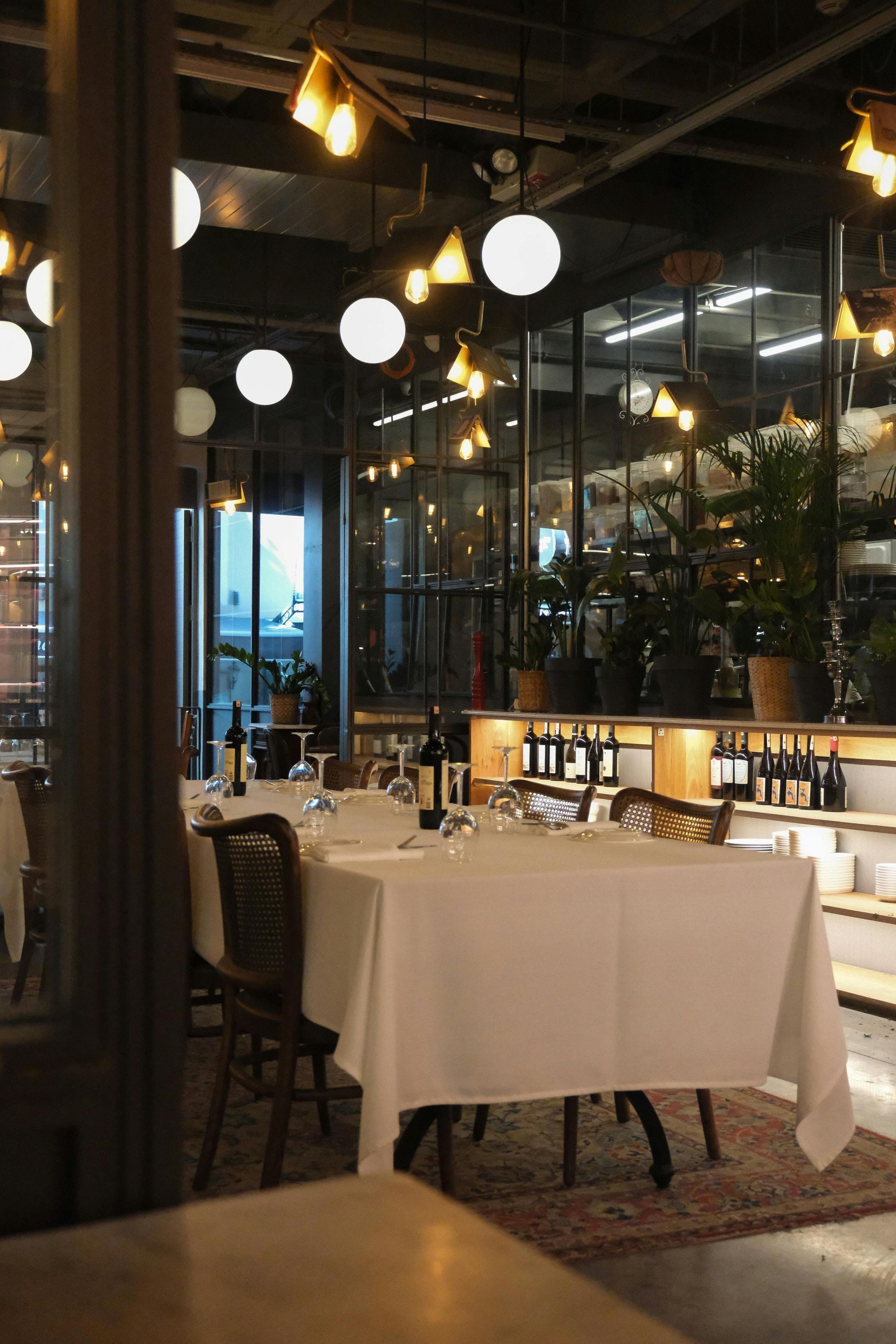 Dimly lit restaurant dining area with white tablecloths, hanging globe lights, and glass walls.