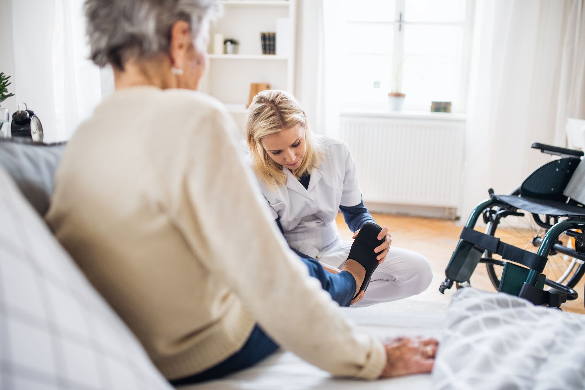 Caregiver helping a seated patient in a bright home living room near a wheelchair