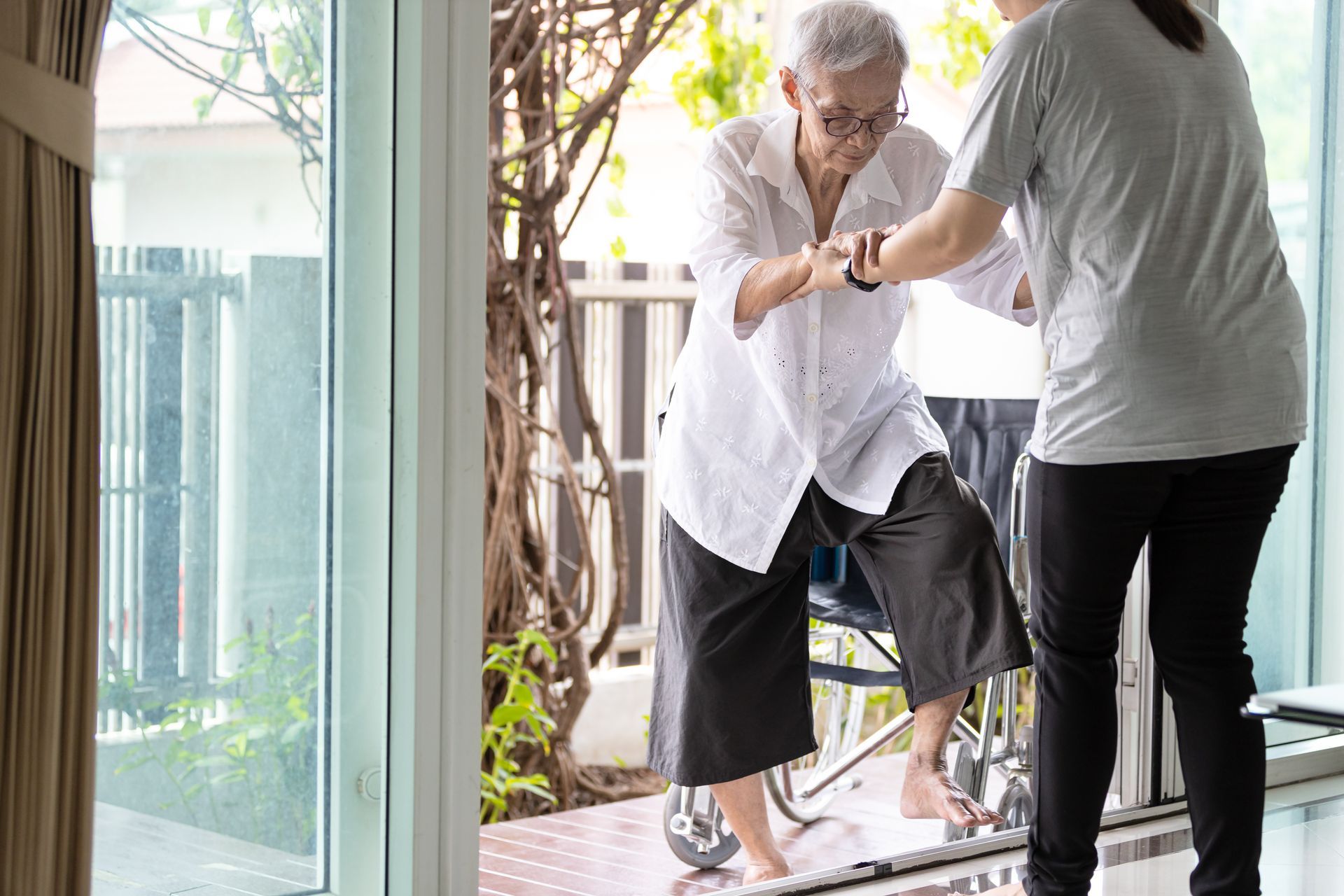 Person helping another step through a doorway indoors, with a cane nearby and bright patio light outside