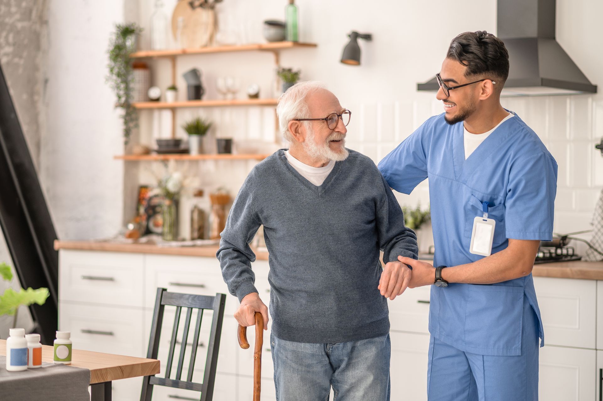 A smiling caregiver, wearing a blue uniform, is supporting a senior with a walking stick.