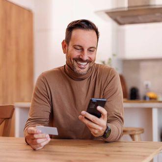 Portrait of happy mid adult man with credit card making online payment over smart phone at dining table