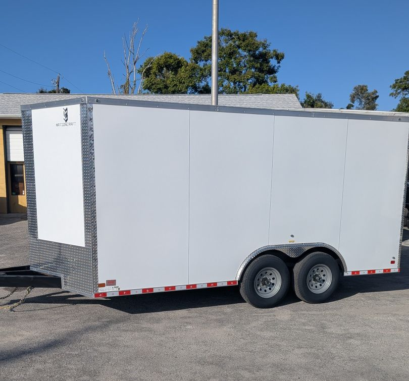 White enclosed dual-axle cargo trailer parked outdoors on pavement under a clear blue sky.