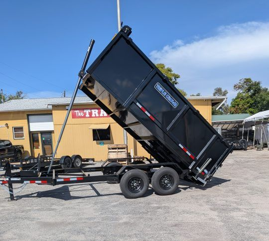 Black dump trailer raised at an angle in a gravel lot beside a yellow building under a blue sky