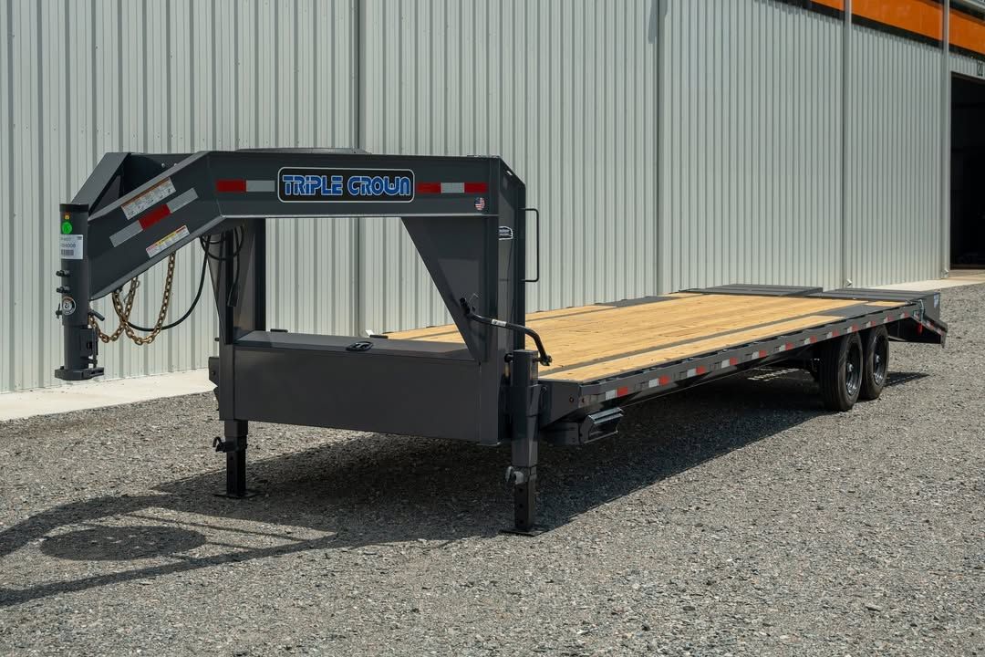 Black utility flatbed trailer with gooseneck hitch on gravel beside a metal building