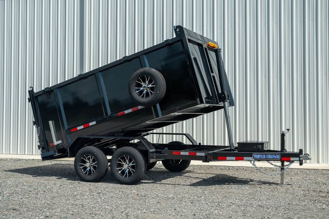 Black dump trailer tipped on its side, leaning on gravel beside a corrugated metal wall.