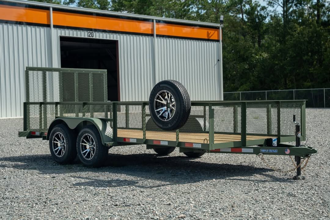 Green utility trailer with mesh sides and two wheels, parked on gravel in front of a metal building.