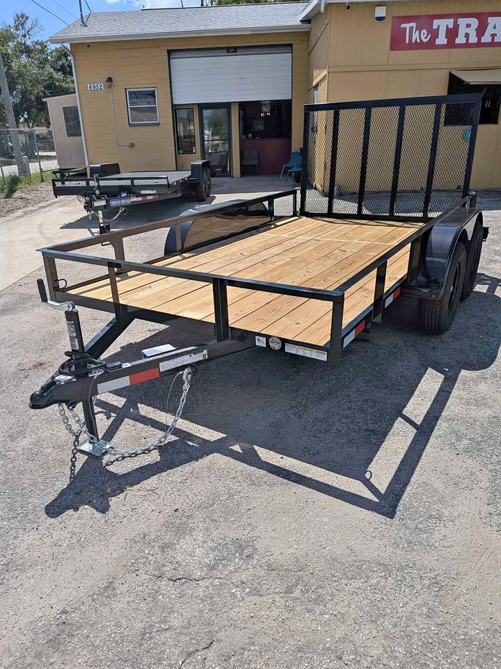 Black utility trailer with wooden floor and mesh ramp, parked outside a building on a sunny day