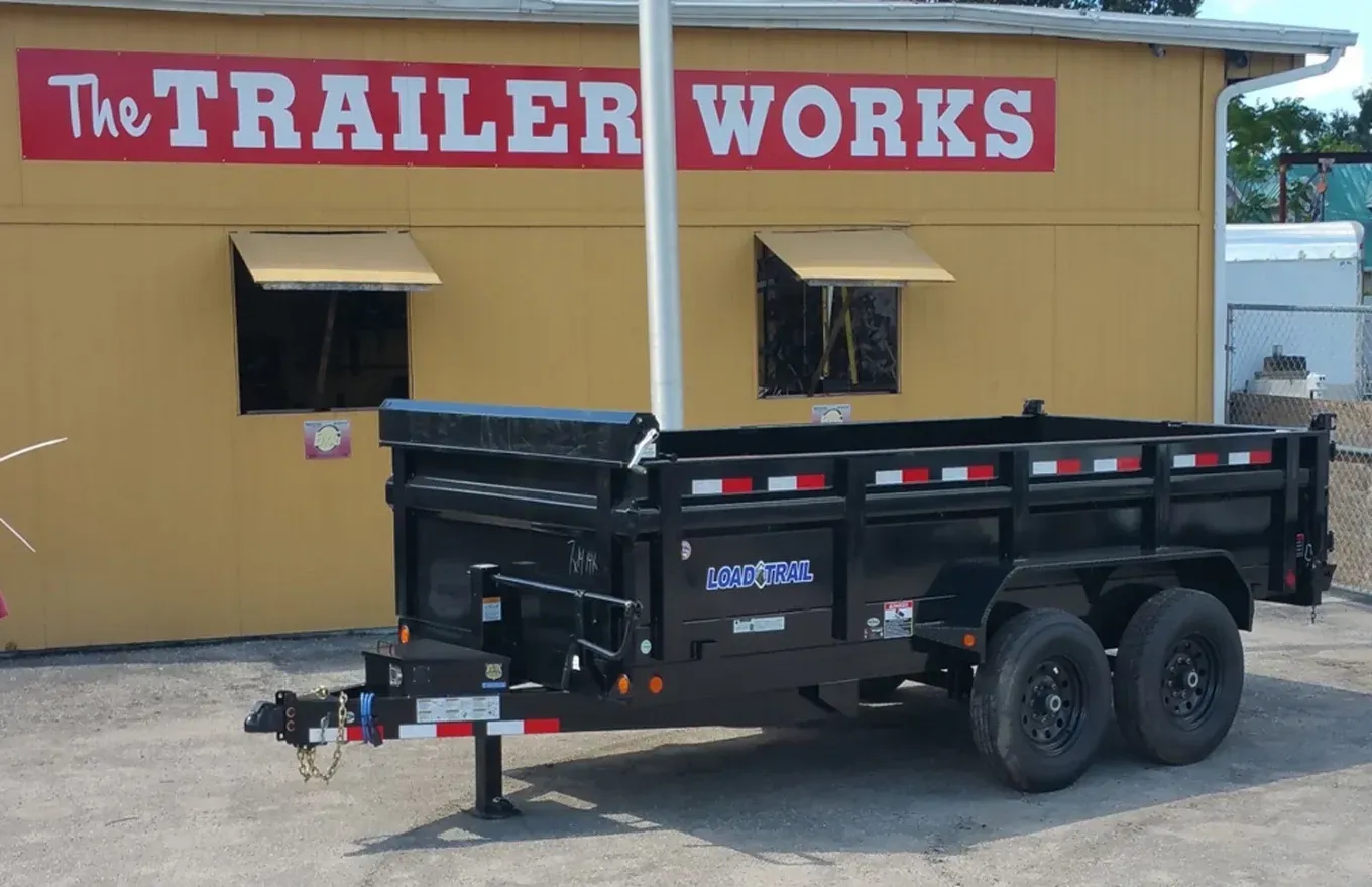 Black dump trailer parked outside The Trailer Works building with yellow walls and red sign.