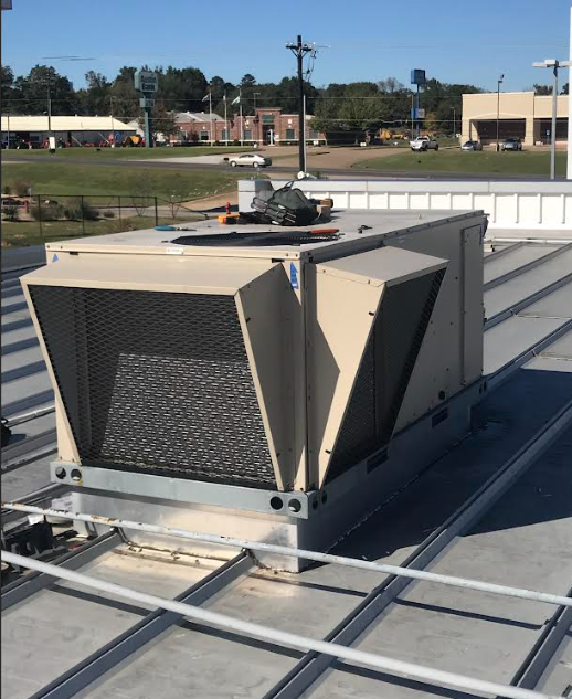 A light gray American Standard commercial HVAC rooftop unit sits on a flat surface beneath a building overhang. A light gray American Standard commercial HVAC rooftop unit sits on a flat surface beneath a building overhang.