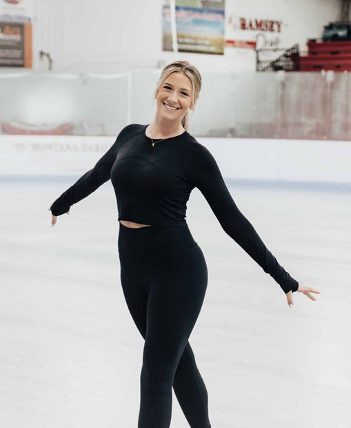 Figure skater in black outfit posing on an indoor ice rink with red stadium seats in the background.