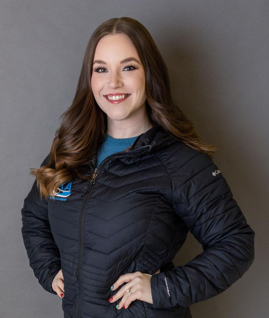 Woman smiles, hands on hips, wearing a black jacket; gray backdrop.