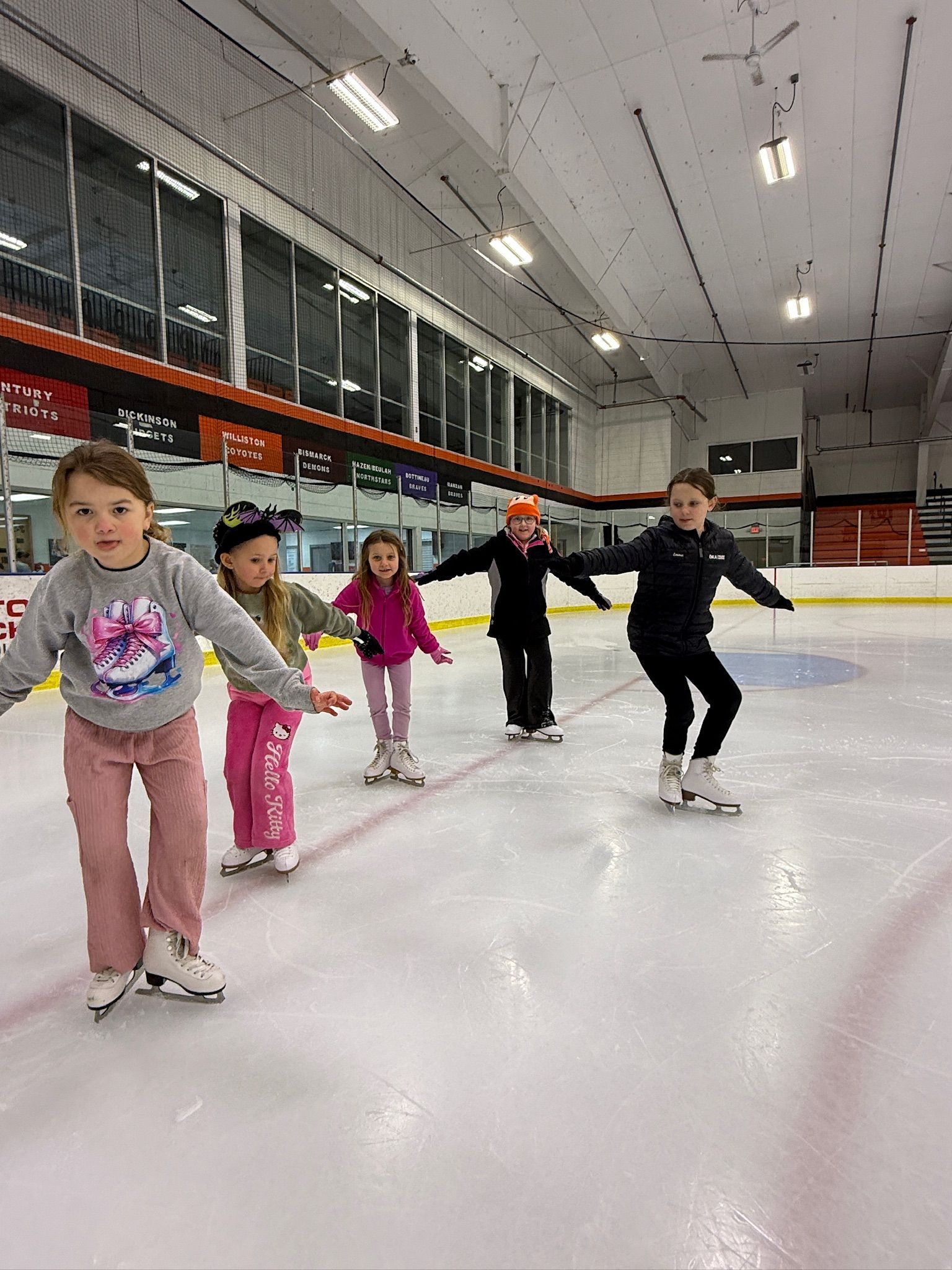 Children holding hands and ice skating in a line on an indoor rink