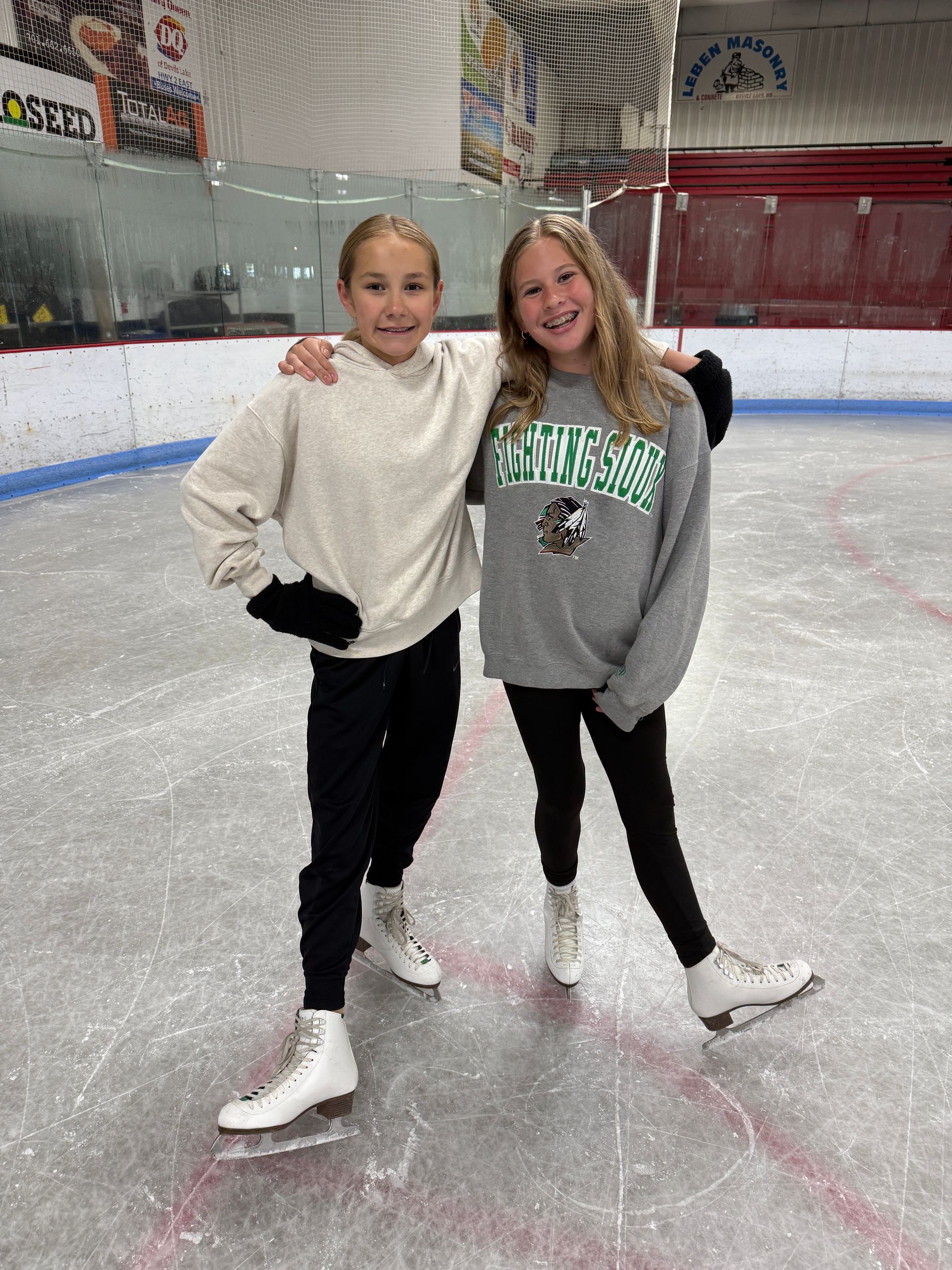 Two people posing on an ice rink, wearing white figure skates and casual winter clothes.