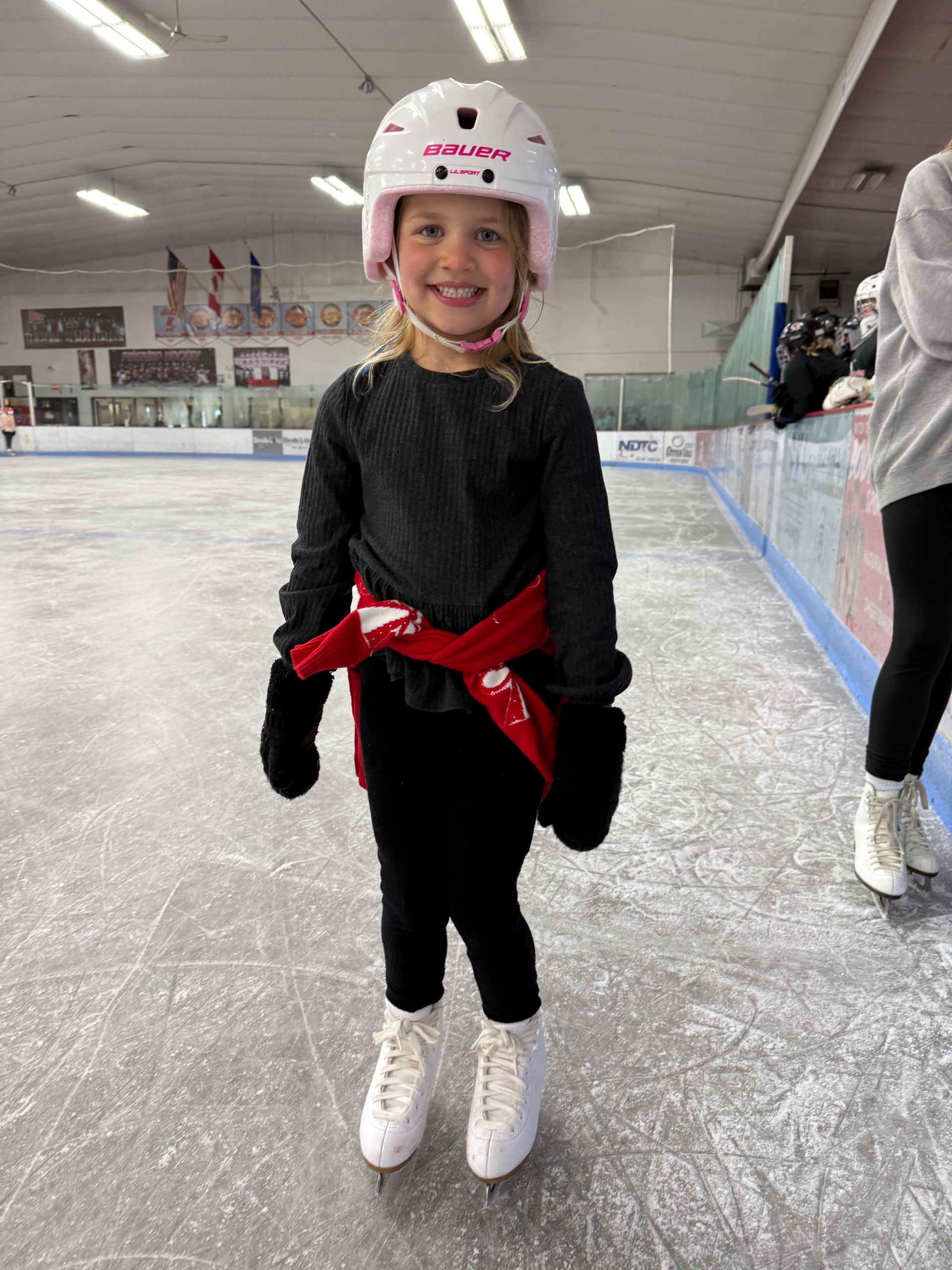 Smiling child in a white helmet and black outfit with red sash, standing on an indoor ice rink
