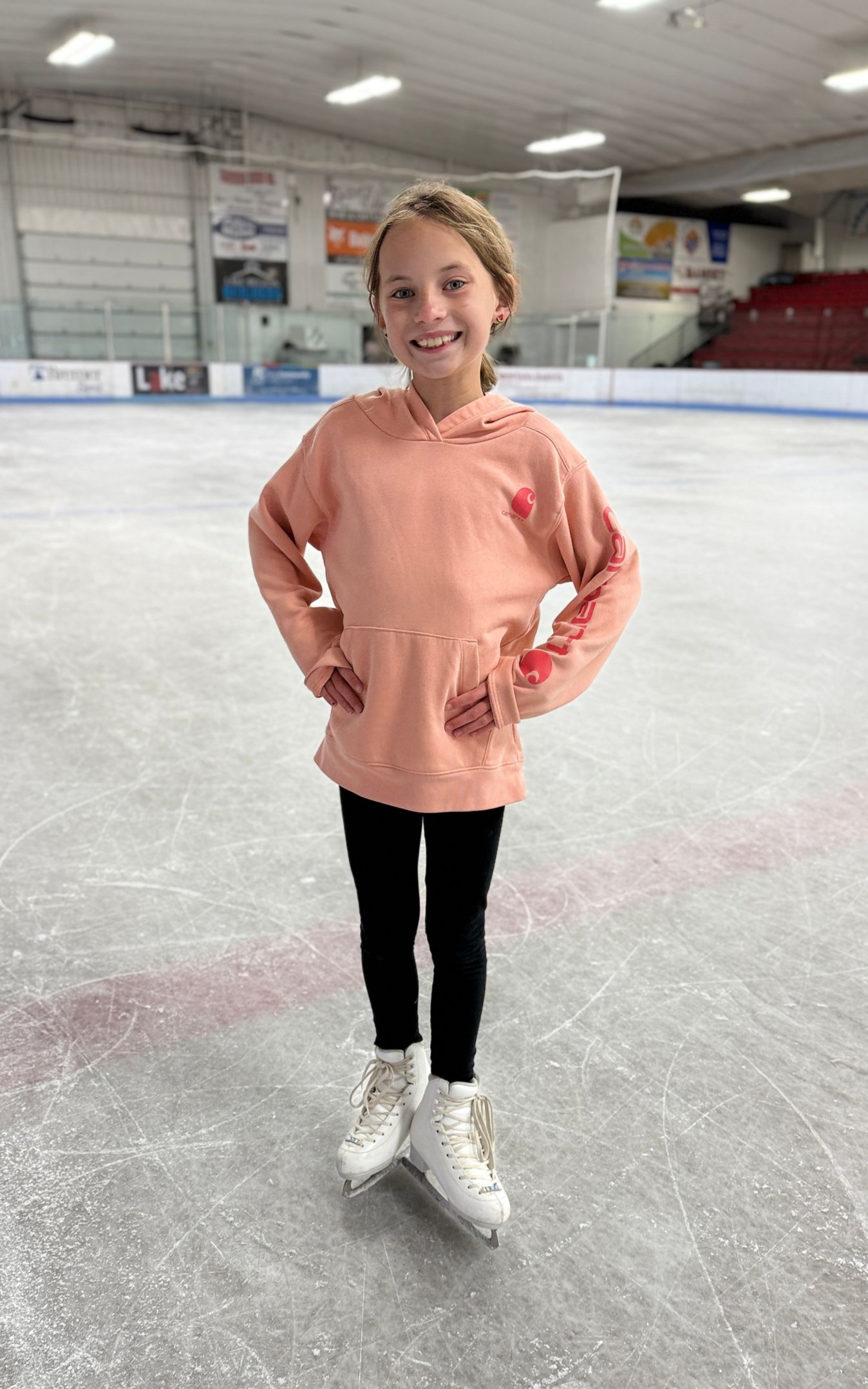 Young figure skater in a peach top and black leggings standing on an indoor ice rink with hands on hips.