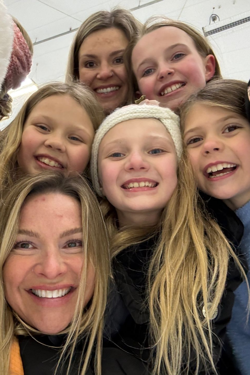 Smiling group selfie of six people indoors, close together against a white wall.