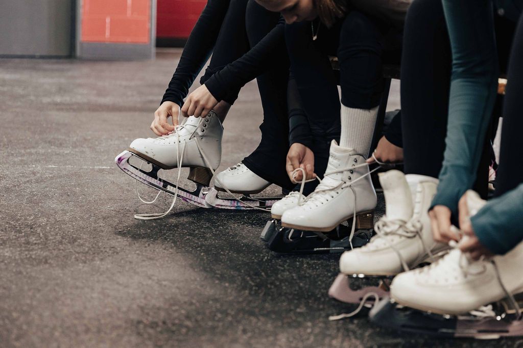 People in black pants are tying white ice skates while sitting.