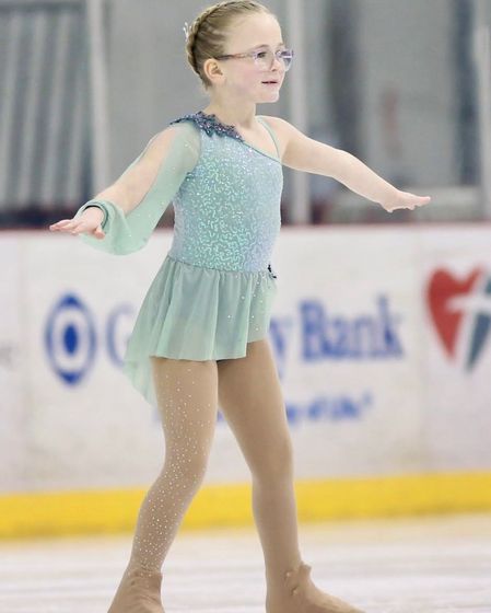 Young figure skater in a teal dress, arms outstretched, skating on ice. The background shows a bank logo.