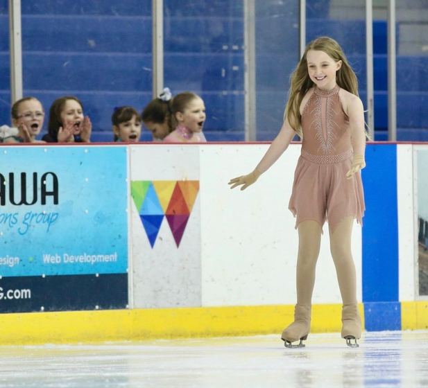 Young ice skater in a pink dress performs. Excited spectators watch behind the rink barrier.