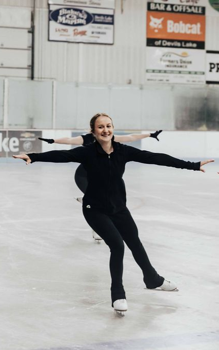 Figure skater in black outfit glides on an indoor ice rink with arms outstretched.