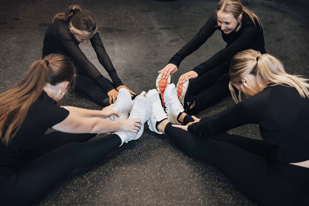 Four people in black stretch a seated partner’s legs on a gym floor.
