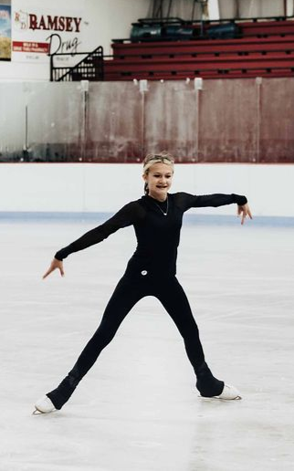 Figure skater in a black outfit gliding on an indoor ice rink with arms extended.