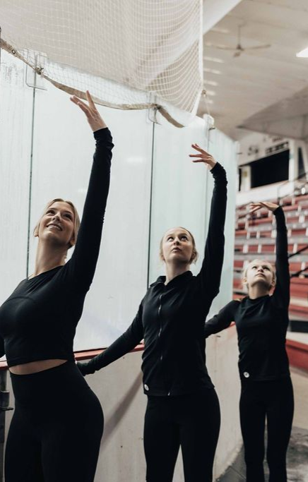 Four dancers in black leotards stretch arms overhead in a rehearsal studio with mirrors and red seating.