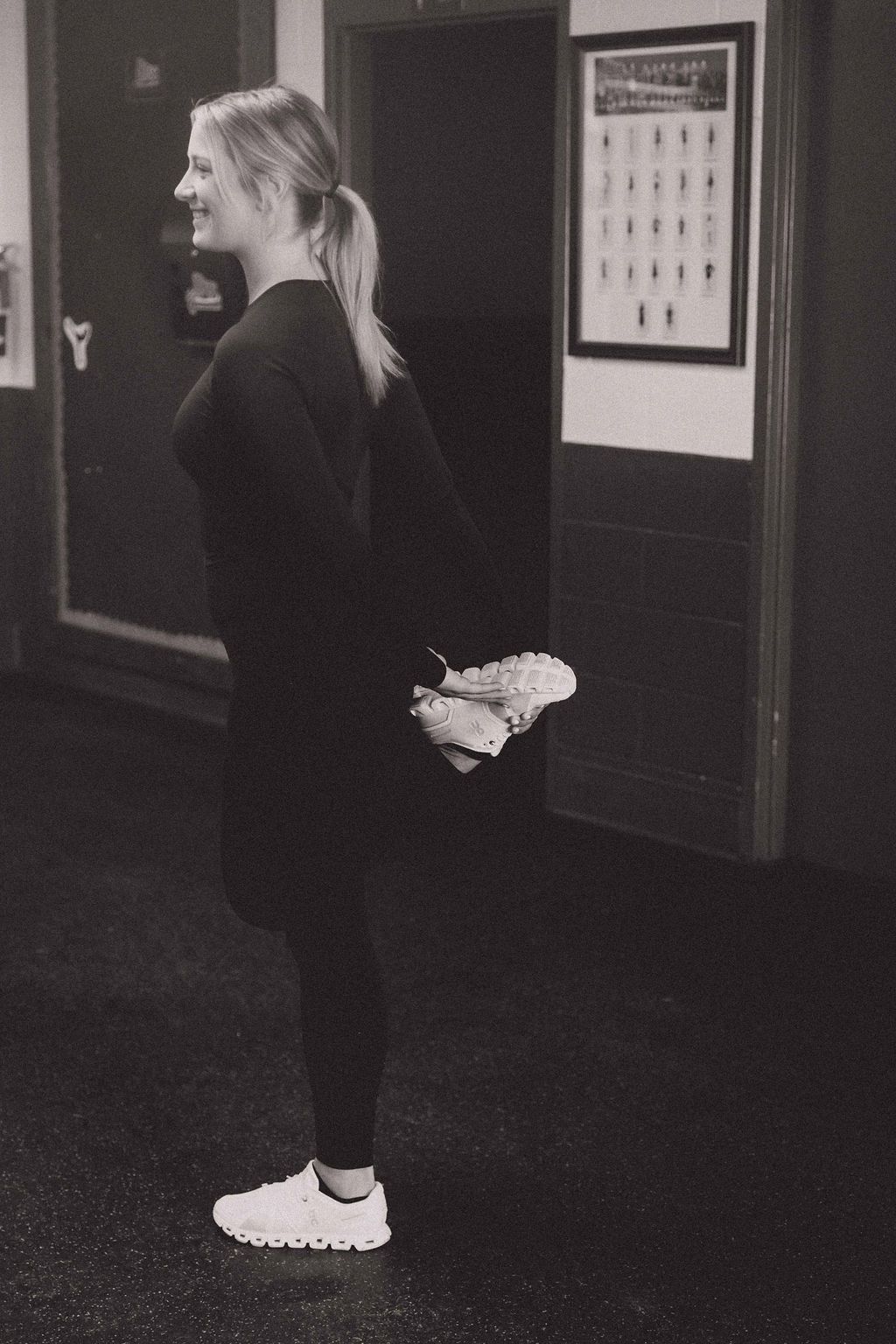 Person standing indoors with hands behind their back, wearing black clothes and white sneakers.
