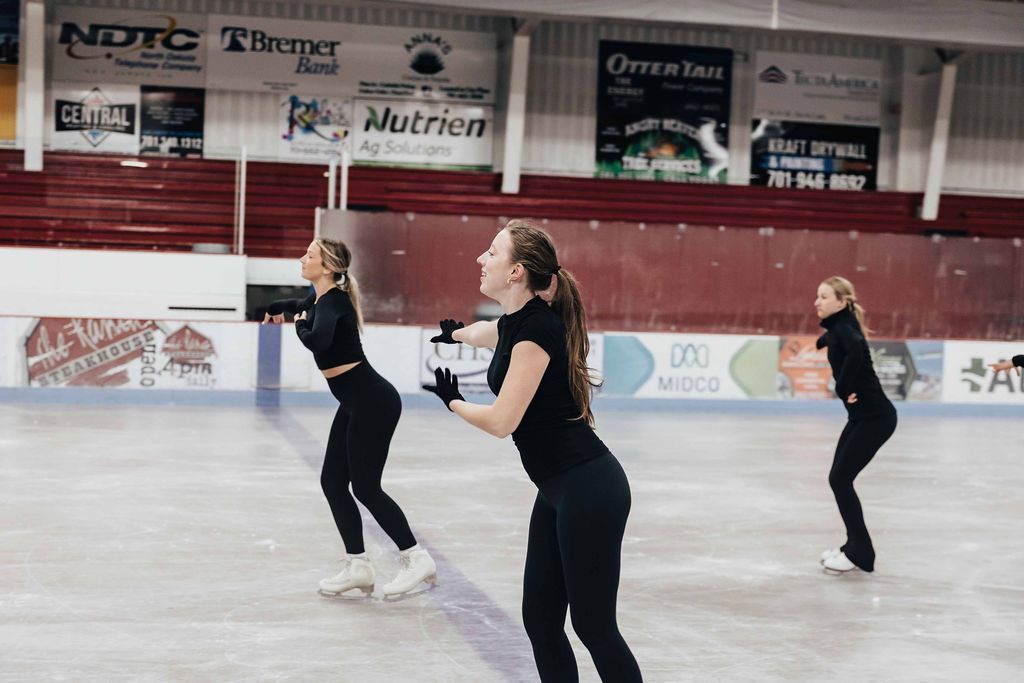 Three skaters in black practice on an indoor ice rink, with banners overhead.