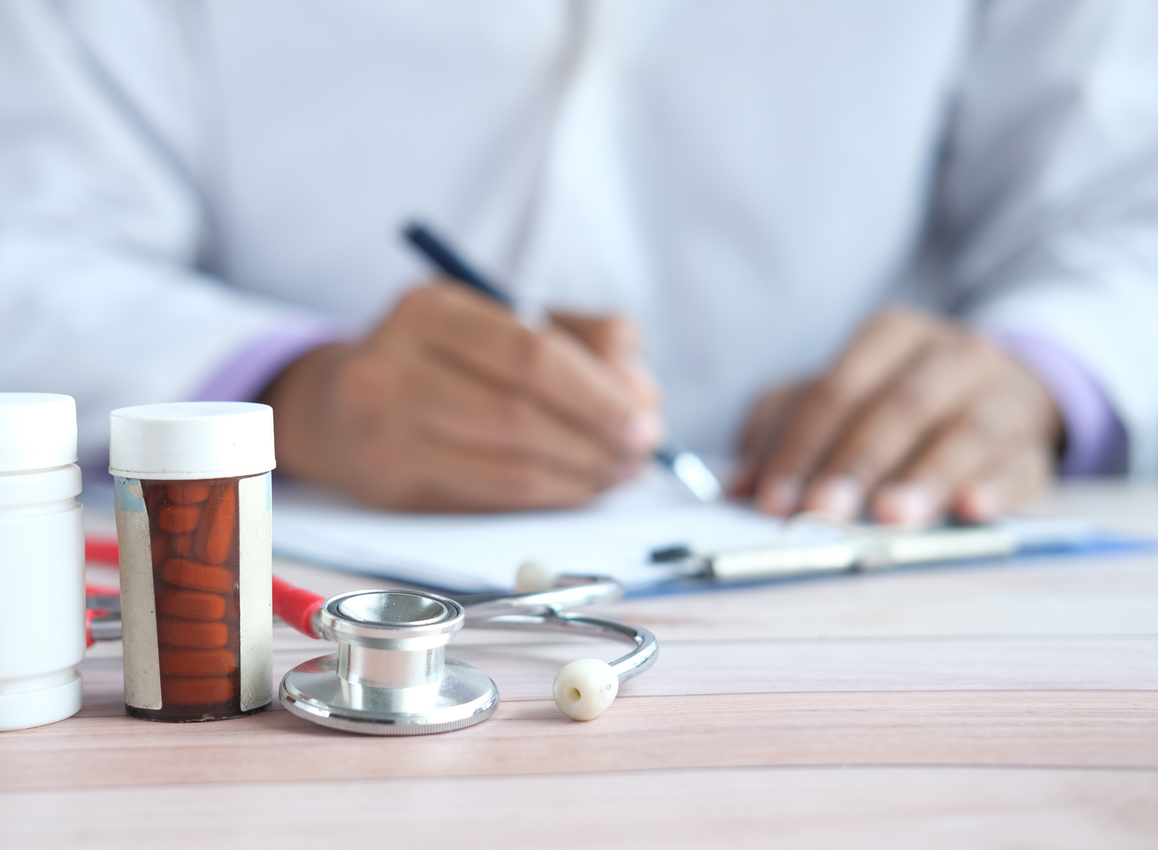 A healthcare professional writing on a clipboard, with a stethoscope and two pill bottles on a desk in the foreground.