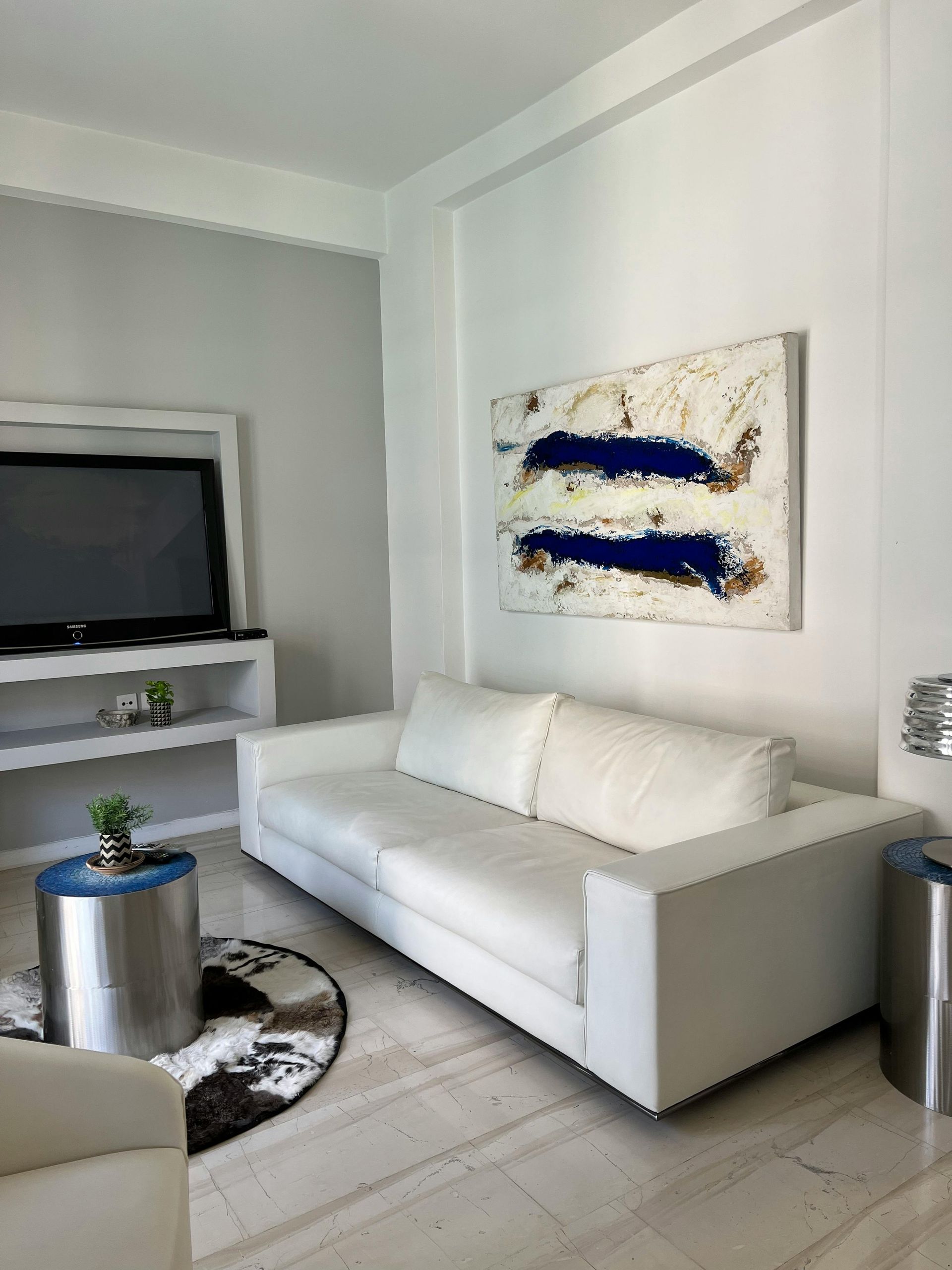 A white sofa sits in a modern living room next to a television, a round metallic table, and an abstract blue-and-white art.