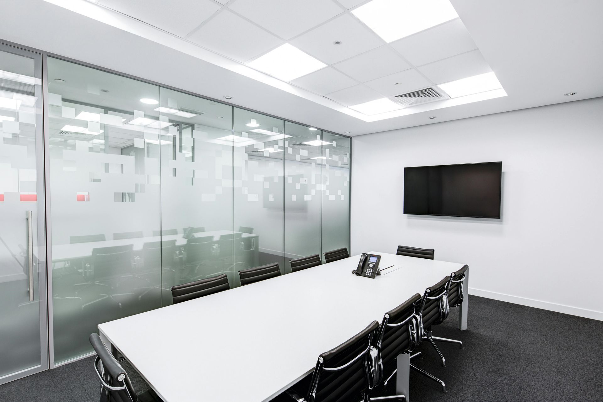 Modern conference room with a white table, black chairs, and a glass-walled partition, set against a plain white wall.