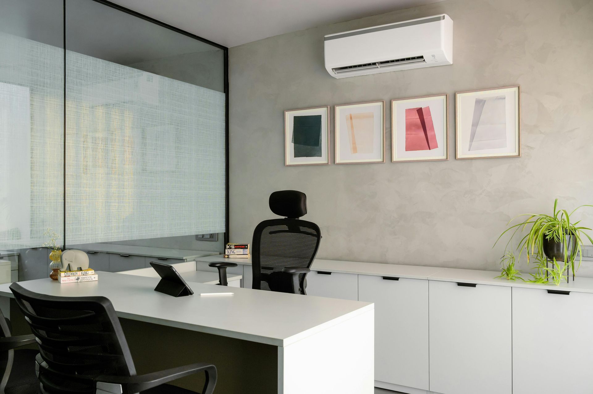 A modern office with a desk, two ergonomic chairs, wall art, a potted plant, and a wall-mounted air conditioner.