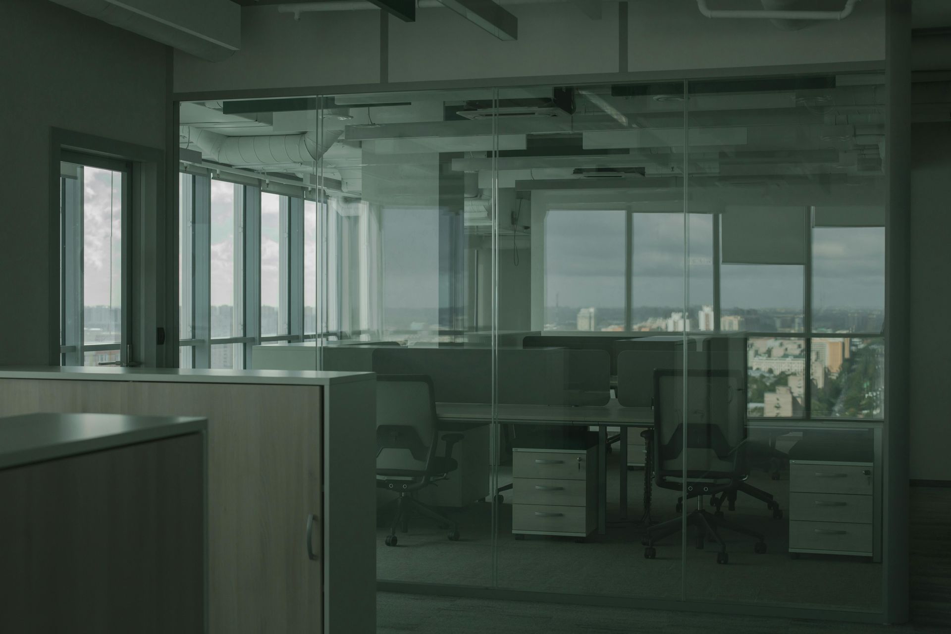 A modern office interior with glass-walled cubicles, desks, and chairs overlooking a city skyline.