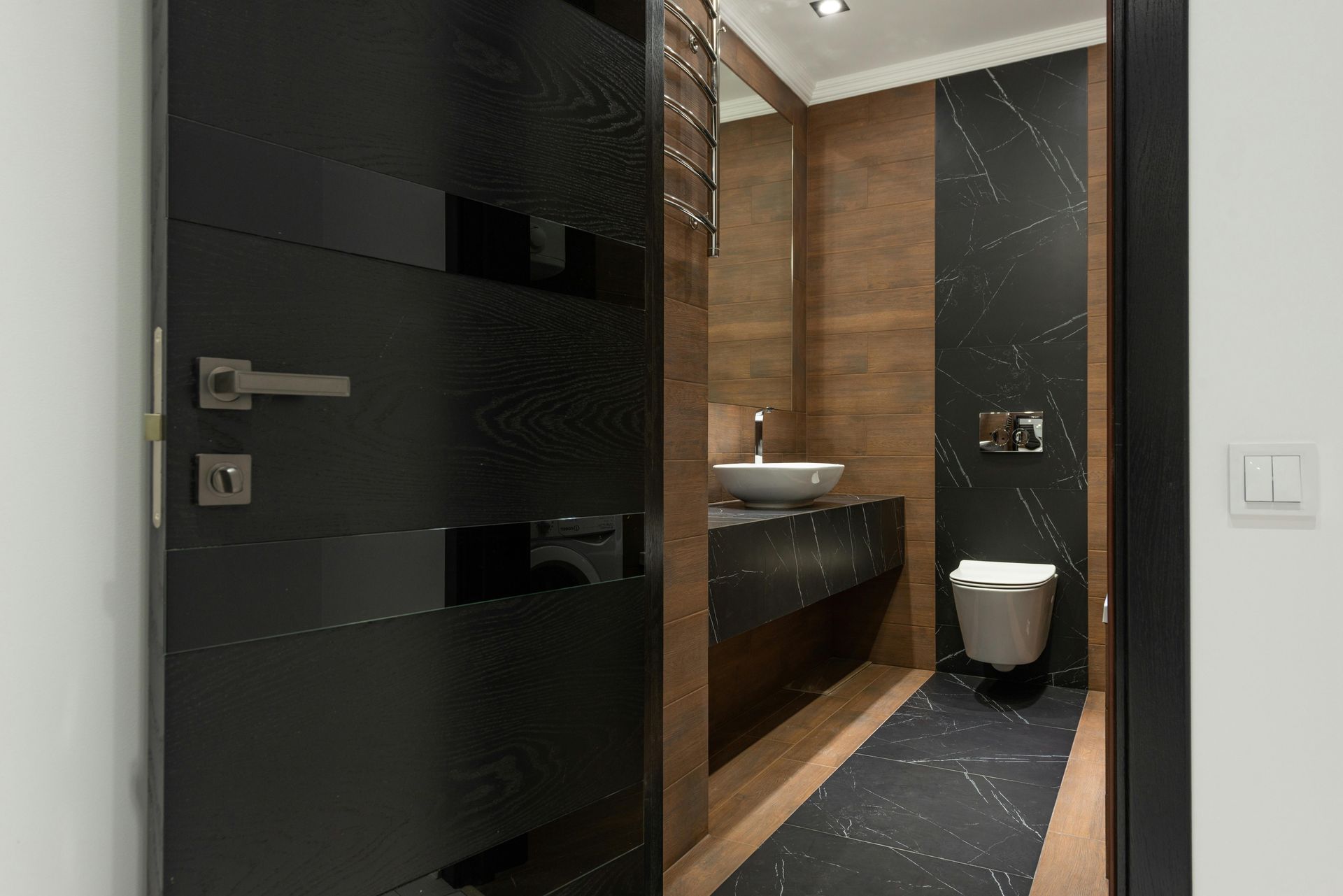 A view through an open dark-paneled door into a modern bathroom with wood-toned walls and black marble accents.