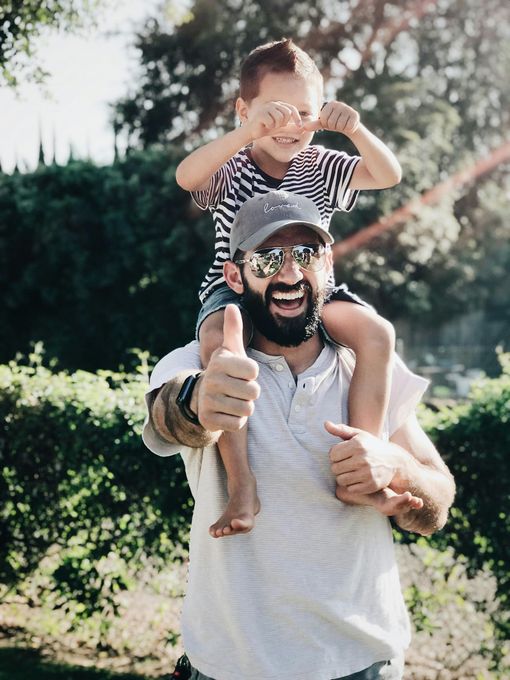 Man carrying a child on his shoulders, both smiling. The man gives a thumbs-up in a sunny outdoor setting.