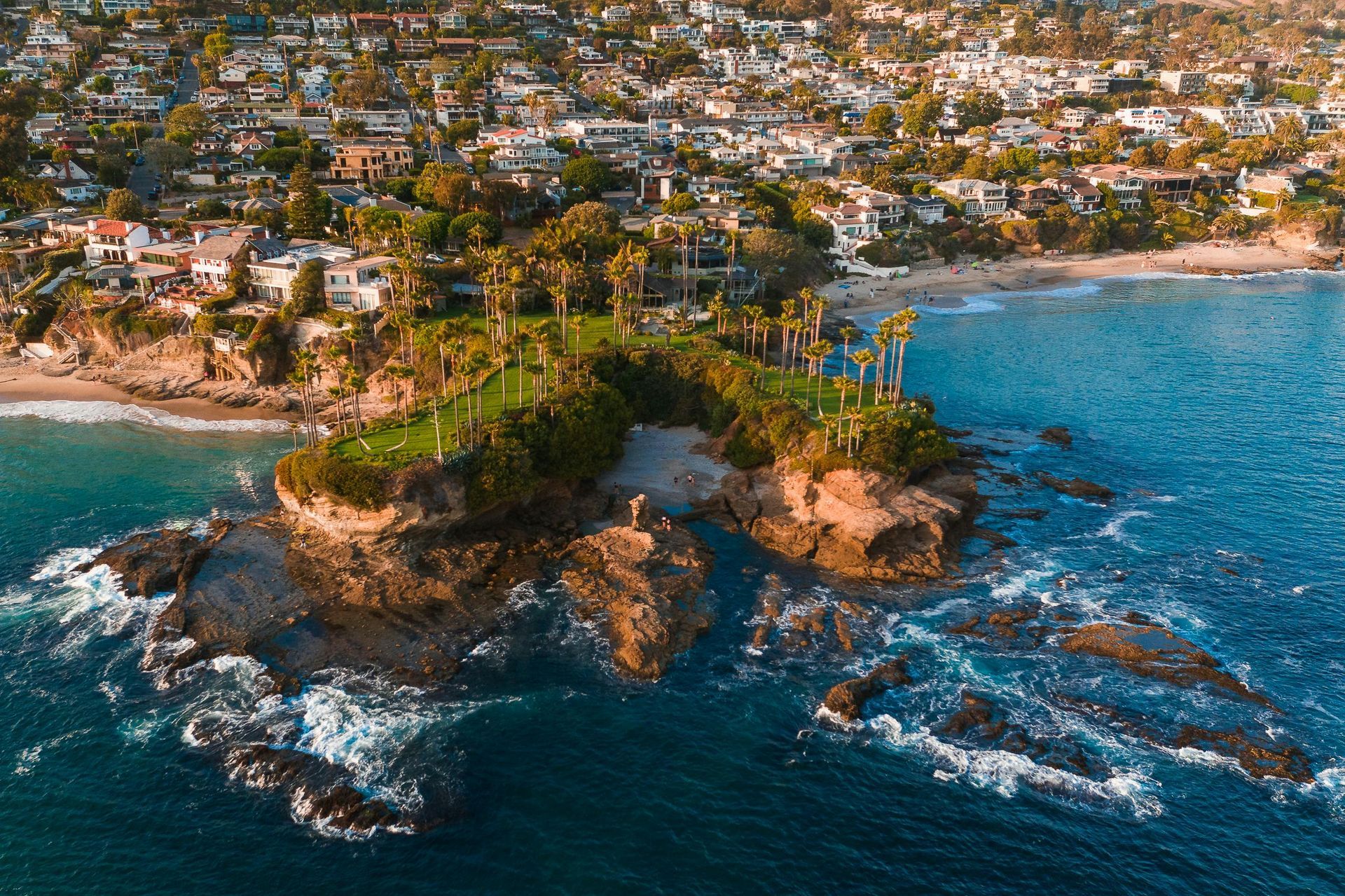Coastal view: rocky outcropping with lush greenery meets ocean; houses dot the coastline under a sunny sky.