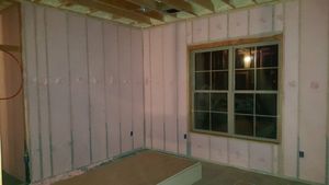 Interior of a room under construction, with pink insulation between wooden studs, a window, and exposed ceiling beams.