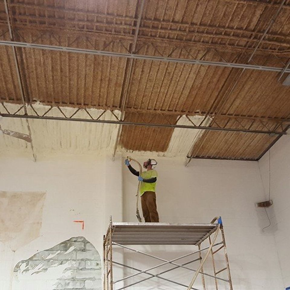 Person spraying insulation on a ceiling, standing on a scaffolding inside a building.