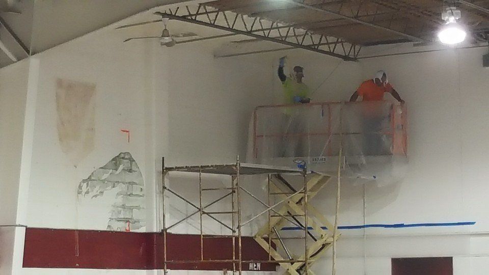 Two workers on a lift, sanding a gymnasium wall with visible dust.