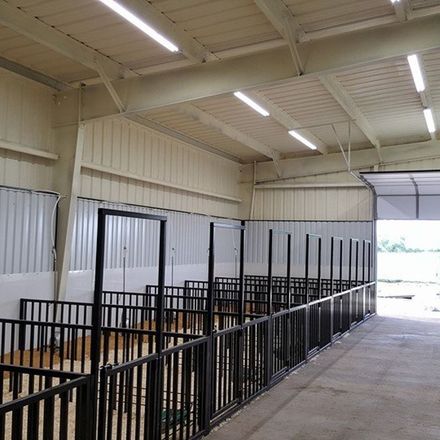 Interior of a beige and white barn with horse stalls, metal framework, and fluorescent lighting.