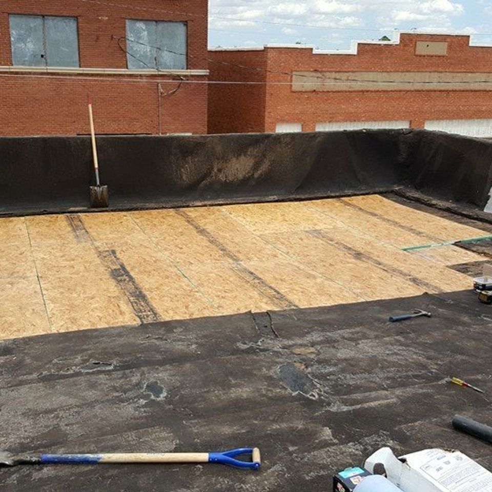 Flat commercial roof with exposed wood and tar, tools scattered. Brick building in background.