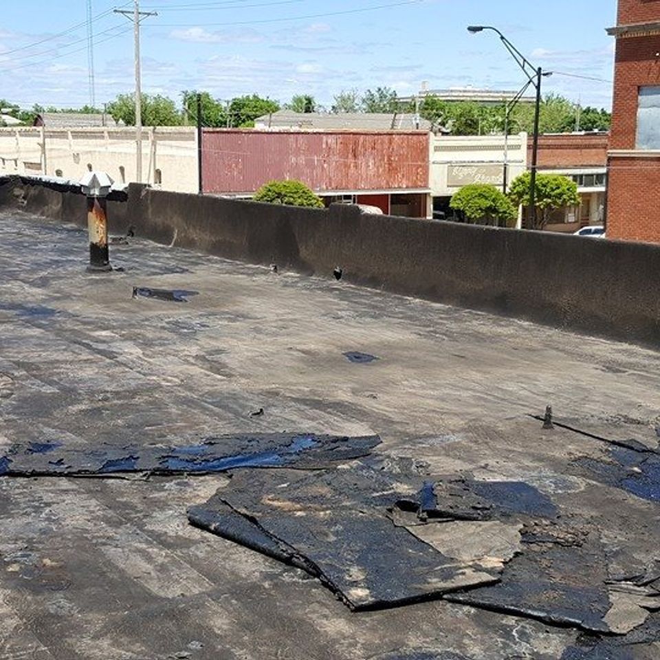 Damaged flat roof with peeling black asphalt against a backdrop of buildings and a bright blue sky.