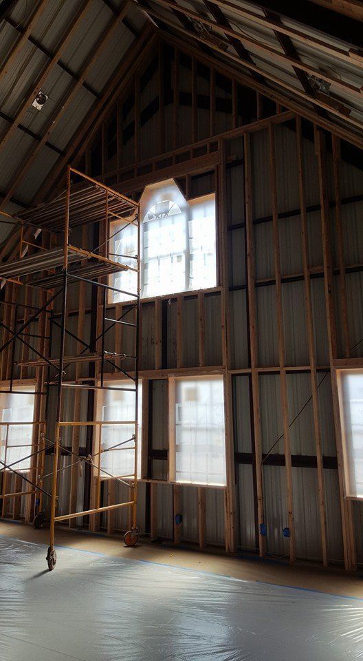 Interior of a building with scaffolding, windows, and exposed wooden framing. The floor appears to be freshly poured concrete.