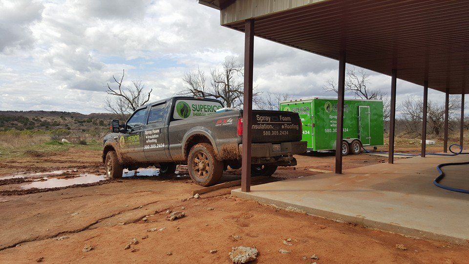 Muddy truck parked by a green trailer under a roofed structure in a dirt area.