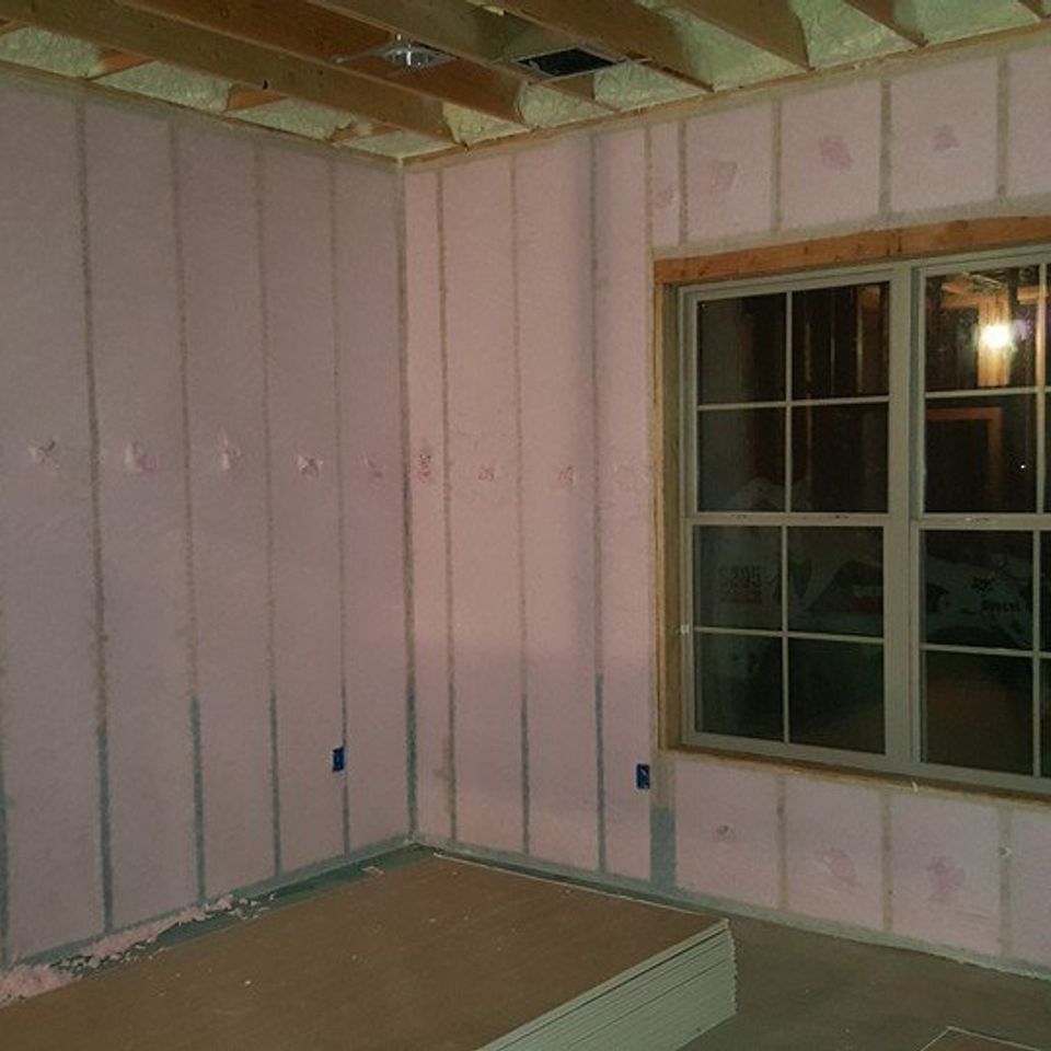 Interior of a room under construction, insulated pink walls, a window, and exposed wooden ceiling beams.
